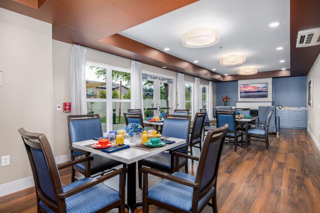 A dining area with wooden flooring, featuring several tables set with dishes, drinks, and flowers. Large windows allow natural light, and comfortable chairs surround the tables. A painting is visible on the wall at the far end.